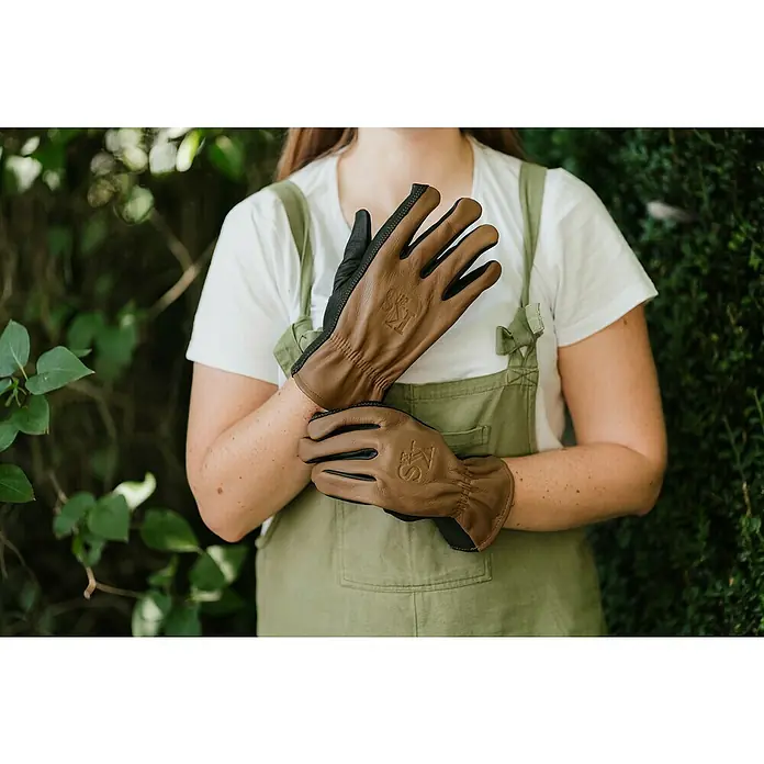 Kent & Stowe Gartenhandschuhe Sure & Soft - S, Braun/Schwarz - Nahaufnahme einer Person, die hochwertige Ledergartenhandschuhe in Braun und Schwarz mit aufgesticktem Logo trägt. Perfekt für Gartenarbeiten!