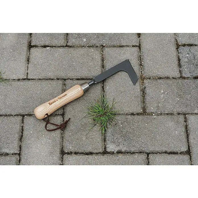 Kent & Stowe Fugenkratzer Carbon Steel - Stahl, Kurzstiel - Nahaufnahme eines Kent & Stowe Fugenkratzers mit Holzgriff, der neben einem Grasbüschel auf Pflastersteinen liegt.