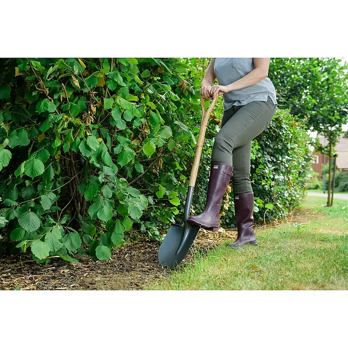 Kent & Stowe Schaufel - Breite: 22 cm, Anthrazit/Holz - Eine Person, die einen Spaten der Marke Kirk Stows in einem Garten in der Nähe einer Hecke mit grünen Blättern hält.