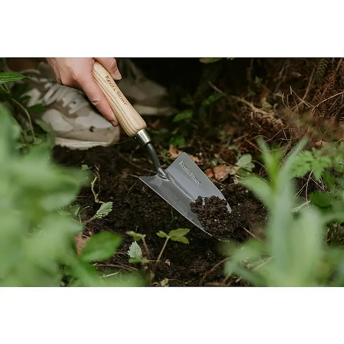 Kent & Stowe Pflanzkelle Carbon Steel - 33,5 cm - Nahaufnahme eines Kent & Stowe Spatens im Einsatz, der Erde aushebt, mit Fokus auf die Marke und die Funktionalität des Werkzeugs.
