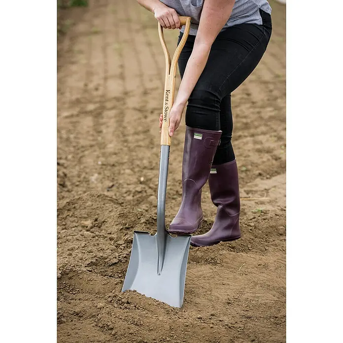 Kent & Stowe Schaufel - Breite: 24 cm, Natur/ Anthrazit - Nahaufnahme einer Person, die einen Kent & Stowe Spaten in einen Ackerboden sticht, ideal für Gartenarbeiten.