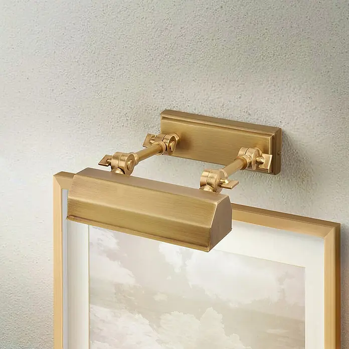Elstead LED-Bilderleuchte Chawton - 40 W, L x B x H: 18,6 x 18,2 x 11,9 cm, Altmessing - Nahaufnahme einer goldenen Bilderleuchte, die ein gerahmtes Bild beleuchtet, montiert an einer strukturierten, neutralen Wand.