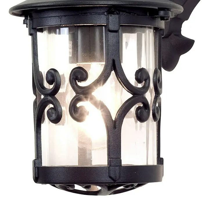 Elstead Außenwandleuchte Hereford - 60 W, 21 x 31 x 31 cm, Schwarz, IP23 - Nahaufnahme einer schwarzen Außenwandleuchte mit dekorativen Verzierungen und klarem Glas, die ein warmes Licht ausstrahlt.