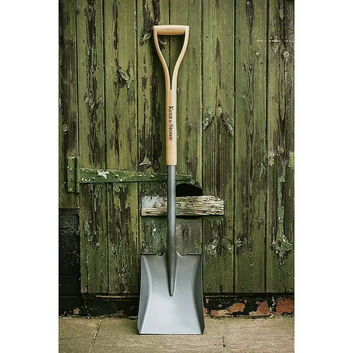 Kent & Stowe Schaufel - Breite: 24 cm, Natur/ Anthrazit - Ein brandneuer Kent & Stowe Spaten lehnt an einer grünen Holzwand, bereit für den Einsatz im Garten. Das Werkzeug mit Holzgriff ist robust und langlebig.