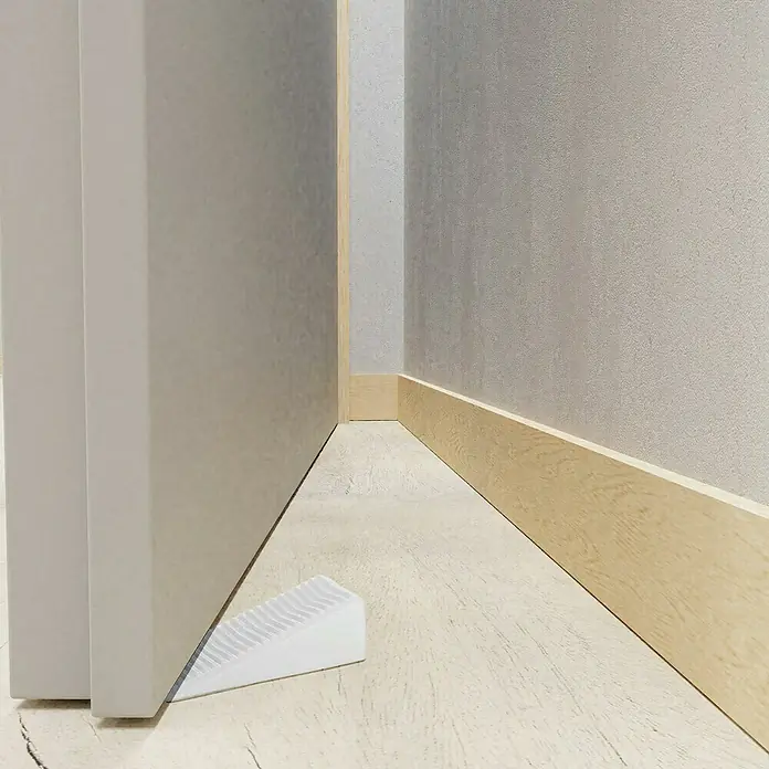 Walteco Türkeil - L x B x H: 15,5 x 50 x 19 mm, Weiß, 4 Stk. - Ein weißer Türstopper aus Kunststoff sichert eine graue Tür auf einem hellen Holzboden, ideal für ein modernes Interieur.