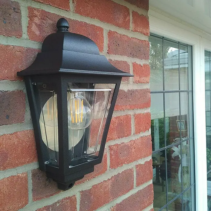 Elstead Außenwandleuchte Deco lane - 60 W, 11 x 32,5 x 32,5 cm, Schwarz, IP44 - Nahaufnahme einer schwarzen Außenwandleuchte mit klarer Glühbirne an einer roten Backsteinmauer neben einem Fenster.