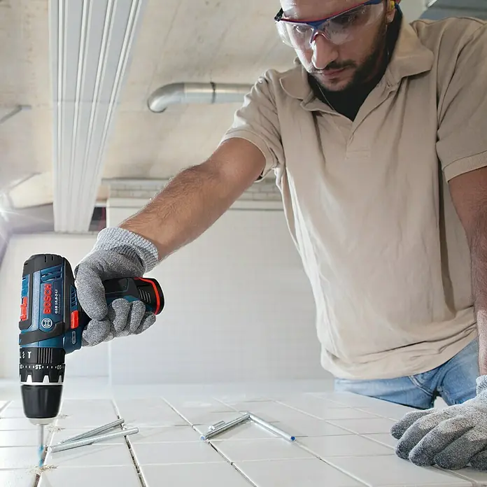 Bosch Professional Akku-Schlagbohrschrauber GSB 12V-15 - 12 V, Ohne Akku, 30 Nm, Ohne Akku, in Karton - Ein Handwerker mit Schutzbrille bohrt mit einer Bosch GSB 10,8-2-LI Bohrmaschine in weiße Fliesen, um sie zu verlegen.
