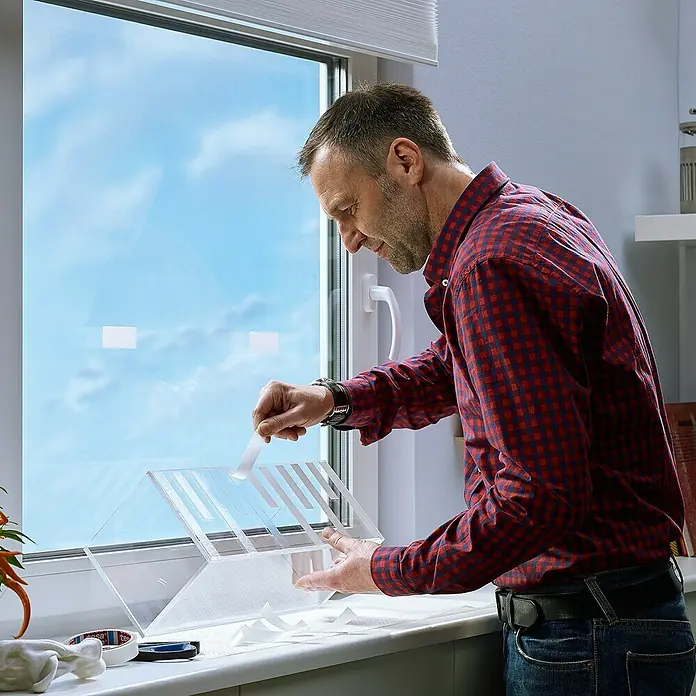 Tesa Montageband - Geeignet für: Glas, Belastbarkeit pro m: 20 kg, 1,5 m x 19 mm - Ein Mann befestigt mit Klebeband ein transparentes Vogelhaus an einem Fenster, um Vögel zu beobachten und die Natur aus nächster Nähe zu erleben.