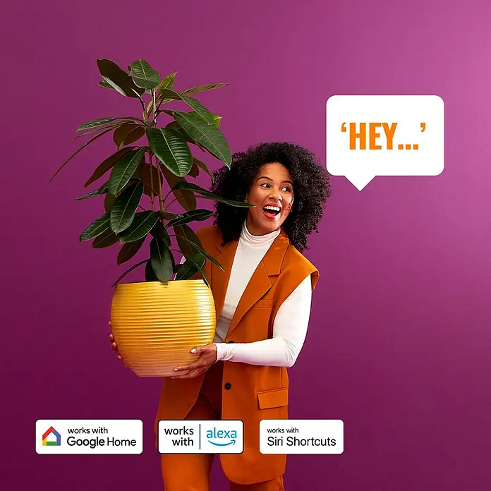 Philips Wiz Smart-LED-Lampe - E27, Dimmbar, Mehrfarbig, 806 lm, 6,7 W, G125 - Eine Frau hält eine Zimmerpflanze und eine Sprechblase mit "HEY..." daneben. Das Produkt ist kompatibel mit Google Home, Alexa und Siri Shortcuts.