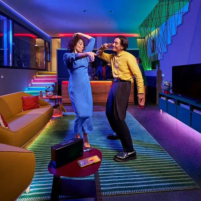 Philips Wiz Smart-LED-Band - Länge: 20 m, RGBW, 36 W, 630 lm - Eine schwarze Box und ein Smartphone stehen auf einem Tisch in einem Raum mit tanzenden Menschen, der mit farbigen Lichtern beleuchtet ist.
