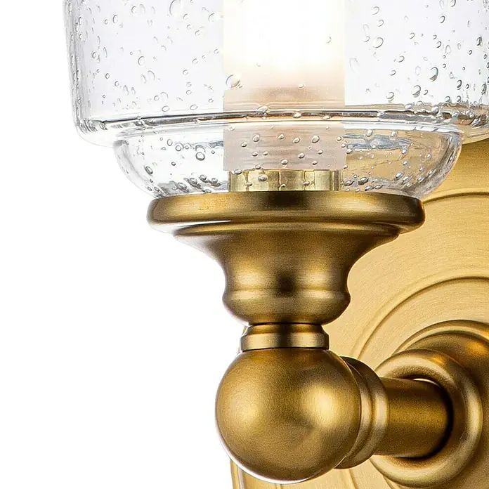 Elstead LED-Wandleuchte Hugenot - 3 W, L x B x H: 15,2 x 13,3 x 24,8 cm, Messing, Warmweiß - Nahaufnahme einer goldenen Wandleuchte mit einem transparenten Glasschirm, der ein warmes Licht ausstrahlt.