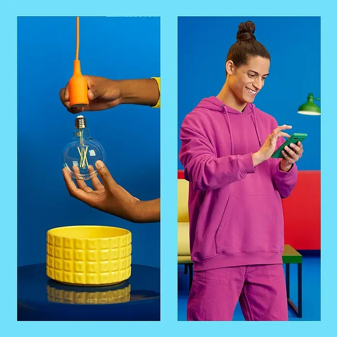 Philips Wiz Smart-LED-Lampe - E27, Dimmbar, Mehrfarbig, 806 lm, 6,7 W, G125 - Das Bild zeigt eine Glühbirne, eine gelbe Keramikschale und einen Mann, der ein grünes Smartphone benutzt. Die Szene ist vor einem blauen Hintergrund.