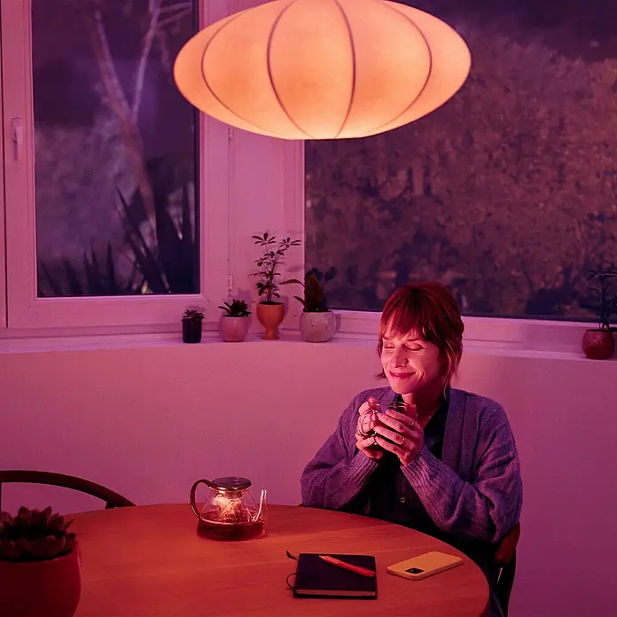 Philips Wiz Smart-LED-Lampe - E27, Dimmbar, RGBW, 12,3 W, 1 Stk. - Frau genießt eine Tasse Tee unter einer stilvollen Lampe, die eine warme Atmosphäre im Raum schafft.