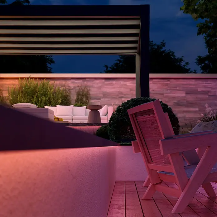 Philips Hue LED-Stripes Outdoor White and Colours Gradient Flux - Länge: 10 m, Lichtfarbe: RGBW, 3.000 lm, 30 W - Einladende Gartenszene mit Loungemöbeln unter einer Pergola, stimmungsvoll beleuchtet, die zum Verweilen einlädt.