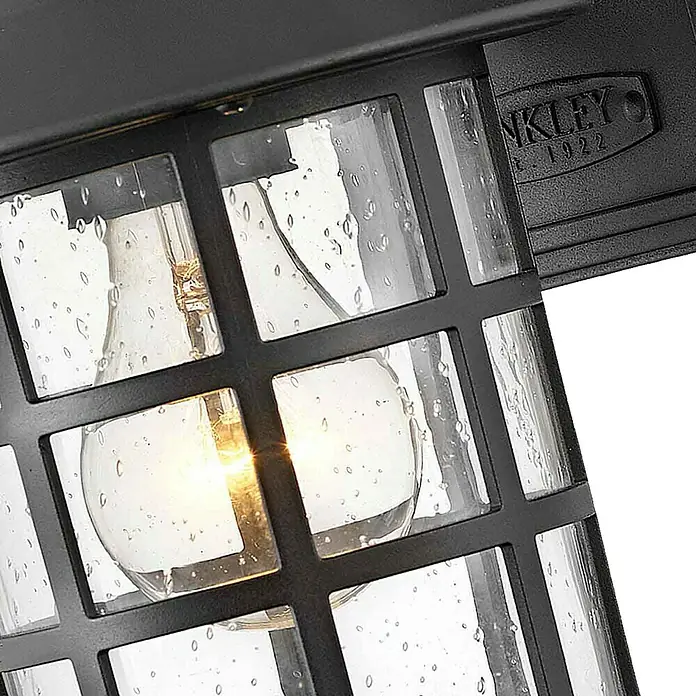 Elstead Außenwandleuchte Freeport - 60 W, 18,5 x 23,8 x 23,8 cm, Schwarz, IP44 - Nahaufnahme einer Außenleuchte mit einem schwarzen Metallrahmen und einer Glühbirne im Inneren, die ein warmes Licht ausstrahlt.