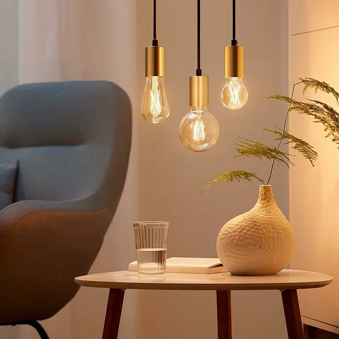 Philips Wiz Smart-LED-Lampe - E27, Dimmbar, Mehrfarbig, 806 lm, 6,7 W, G95 - Drei dekorative Glühbirnen mit goldenen Fassungen hängen über einem Tisch mit Vase, Buch und Glas. Elegante Beleuchtung für ein gemütliches Ambiente.