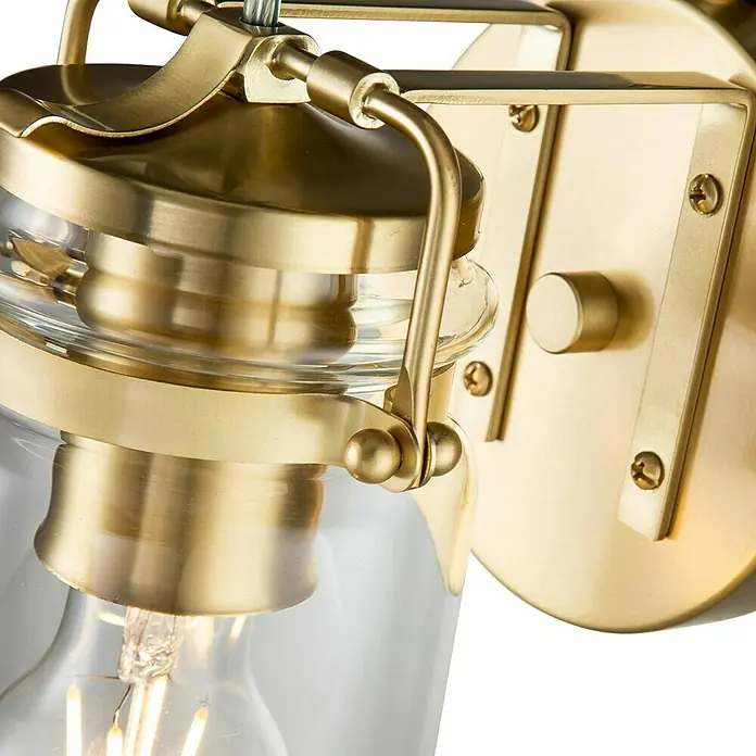 Elstead Wandleuchte Brinley - 60 W, L x B x H: 15,9 x 12,7 x 29,2 cm, Messing, E27 - Nahaufnahme einer goldenen Wandleuchte mit klarem Glasschirm und Edison-Glühbirne, die warmes Licht ausstrahlt.