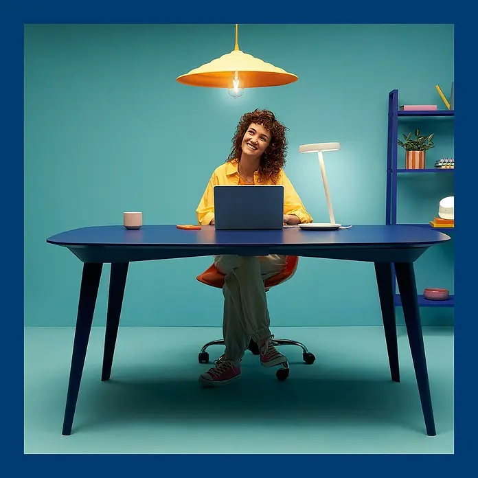 WiZ LED-Tischleuchte Portrait - 10 W, L x B x H: 20,4 x 19,5 x 38 mm, Weiß, Kaltweiß - Eine Frau arbeitet an einem blauen Tisch mit einer Laptop Lampe und einer hängenden Lampe über sich.