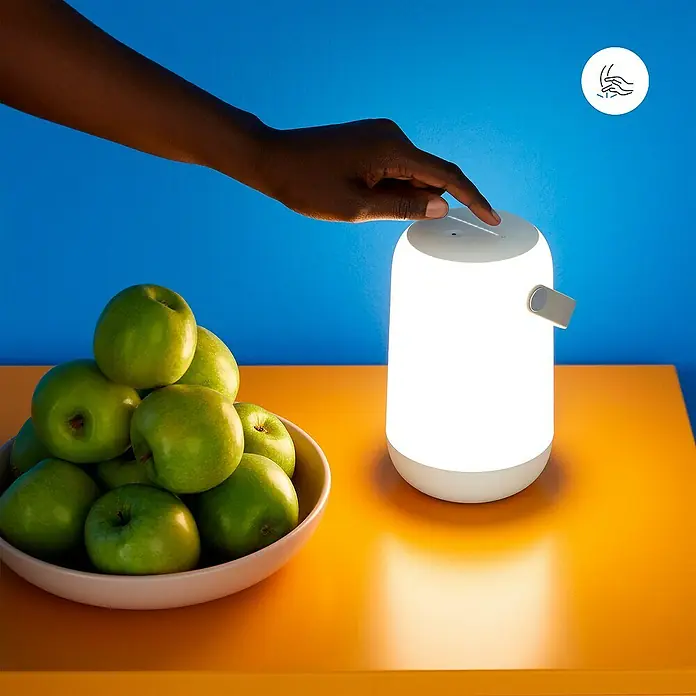 Philips Wiz LED-Tischleuchte Mobile - 14 W, L x B x H: 15 cm x 11,6 cm x 19,3 mm, Weiß, RGB - Nahaufnahme einer Person, die eine tragbare Tischleuchte mit einer Hand berührt, neben einer Schale mit grünen Äpfeln.