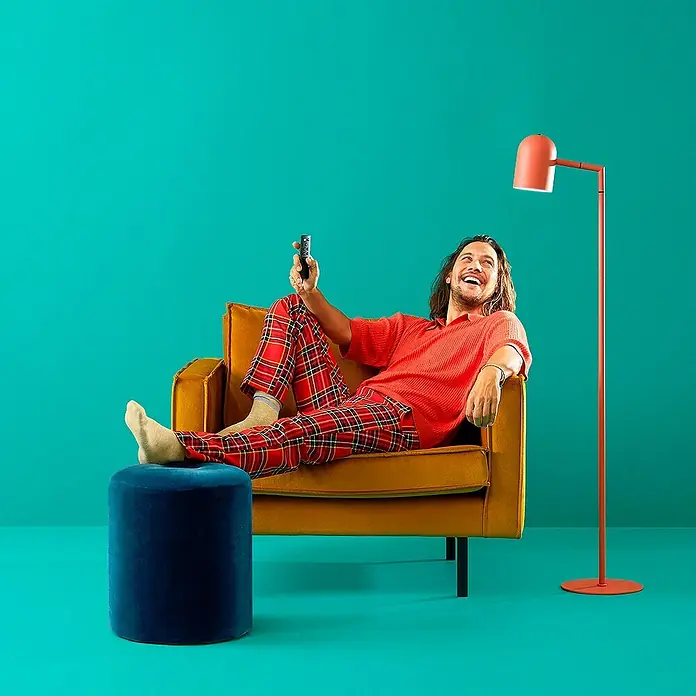 Philips Wiz Smart-Steckdose - Innen, IP20, Weiß, 1 Stk. - Ein Mann entspannt im Sessel mit karierter Hose und Fernbedienung in der Hand. Die Lampe und der Hocker ergänzen das Ensemble farblich.
