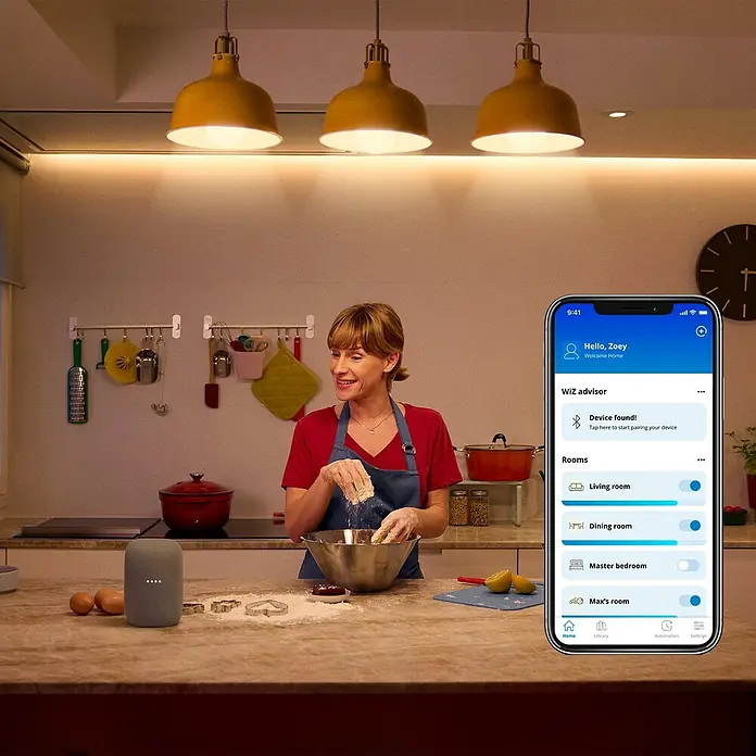 Philips Wiz Smart-LED-Lampe - E27, Dimmbar, RGBW, 12,3 W, 1 Stk. - Intelligente Beleuchtungslösung: Eine Frau backt in der Küche, während sie eine App zur Steuerung der WiZ-Beleuchtung nutzt.