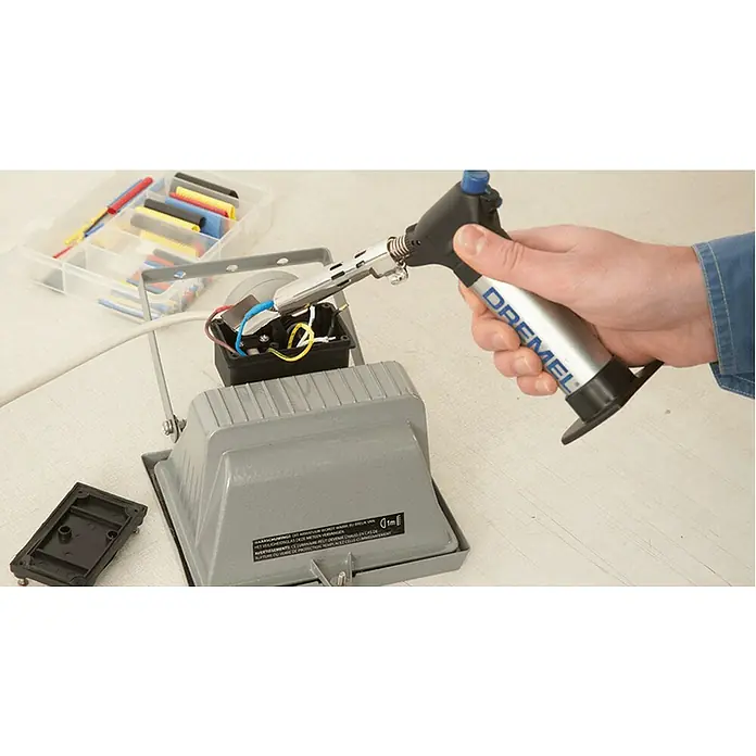 Dremel VersaFlame Hochleistungslötgerät 2200-5 - Geeignet für: Butangas - Ein Dremel-Lötbrenner wird verwendet, um an einer elektrischen Verbindung in einem grauen Gehäuse zu arbeiten. Eine Box mit Schrumpfschläuchen liegt daneben.