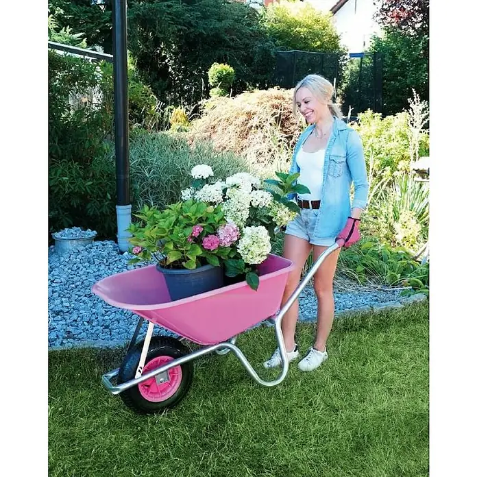 Bauschubkarre "Die Leichte" - 100 l, Traglast: 120 kg, Pink - Eine Frau schiebt eine Schubkarre mit Hortensien durch ihren Garten. Die rosafarbene Schubkarre sticht im Grünen hervor.