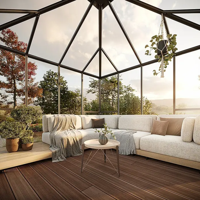 KGT Gewächshaus PaviLux 3,5 - Außenmaß inkl. Dachüberstand (B x T): 350 x 350 cm, 4 mm, Anthrazit-grau, Einscheibensicherheitsglas - Wohnzimmer mit gemütlichem Ecksofa, rundem Tisch und Pflanzen in einem Wintergarten mit Glasdach und Holzboden für entspannte Stunden.