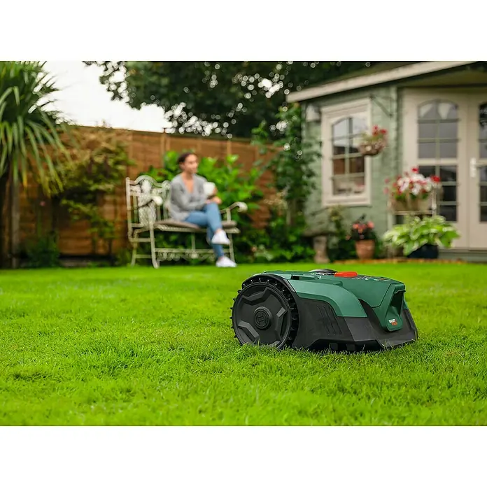 Bosch Power for All 18V Mähroboter VISIMOW 18V-100 - 100 m², Schnittbreite: 16 cm, Kamera-gestützte Navigation, Ultraschallsensorik, Ohne Akku - Der Bosch Mähroboter im Garten, der Rasen ist perfekt gemäht. Im Hintergrund sitzt eine Frau auf einer Bank.