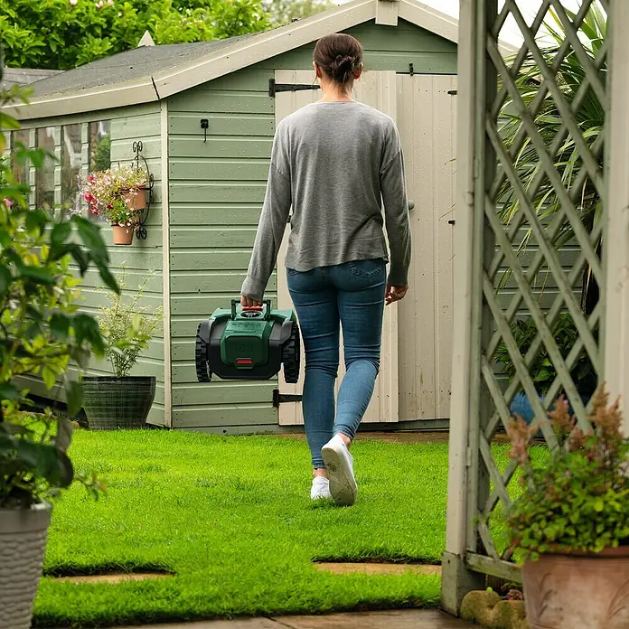 Bosch Power for All 18V Mähroboter VISIMOW 18V-100 - 100 m², Schnittbreite: 16 cm, Kamera-gestützte Navigation, Ultraschallsensorik, Ohne Akku - Frau trägt einen grünen Robotermäher. Ideal zum einfachen Rasenmähen, der Rasenmäher ist kompakt und einfach zu bedienen.