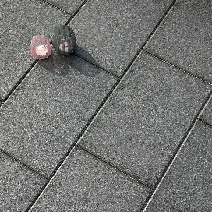 Diephaus Terrassenplatte Redding - 60 x 60 x 4 cm, Schwarz-Basalt, Beton - Graue Pflastersteine mit zwei Kerzenhaltern. Das Bild zeigt die Textur und das Muster der Pflastersteine und die Kerzenhalter als Dekoration.