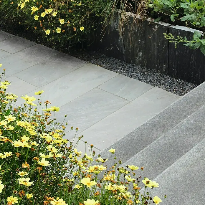 Diephaus Winkelstufe - 80 x 32 x 18 cm, Grau, Beton - Nahaufnahme eines Steingartenbereichs mit gelben Blumen, grauen Steinplatten und dekorativen Steintreppen. Die sorgfältige Gestaltung verbindet Natur und Architektur.