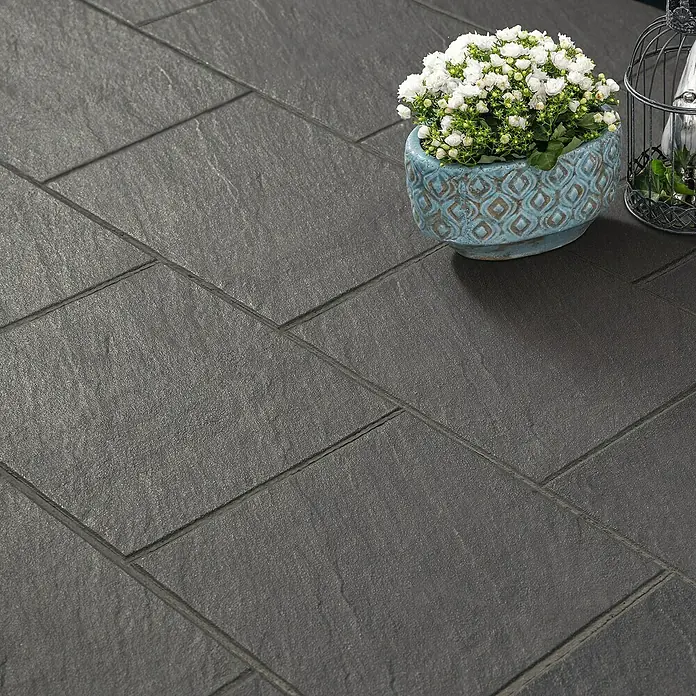 Diephaus Terrassenplatte Rancho - 40 x 40 x 4 cm, Schwarz-Basalt, Beton - Nahaufnahme von dunklen Pflastersteinen mit einer Topfpflanze mit weißen Blumen und einer dekorativen Laterne.
