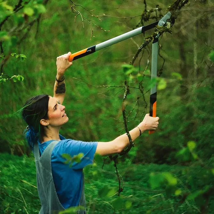 Fiskars Getriebe-Astschere PowerGearX LX98-L - Max. Aststärke: 50 mm, 80 cm - Eine Frau benutzt eine Astschere mit langem Griff, um in einem grünen Wald einen Ast zu schneiden. Das Werkzeug ist grau mit orangefarbenen Akzenten.