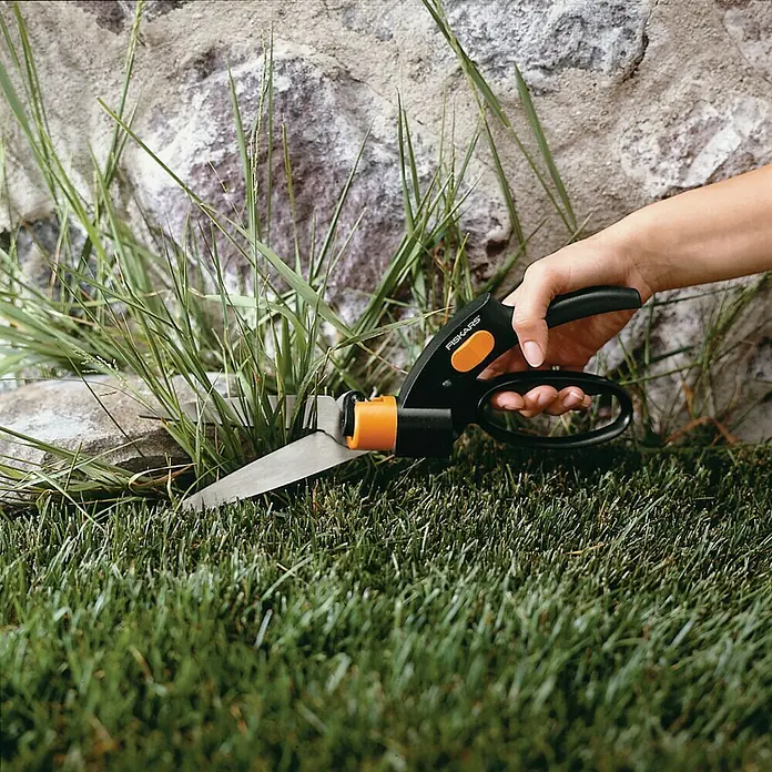 Fiskars Rasenkantenschere Servo System GS42 - Länge: 32 cm - Nahaufnahme einer Hand, die mit einer rotierenden Rasenkantenschere von Fiskars Gras schneidet, vor einer Steinmauer.