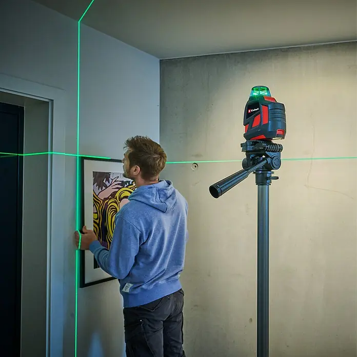 Einhell Adapter TRIPOD - Passend für: Geräte mit einem 1/4" Gewinde - Ein Mann befestigt ein Bild mit der Hilfe eines Einhell Kreuzlinienlasers auf einem Stativ, der grüne Laserlinien an die Wand projiziert.