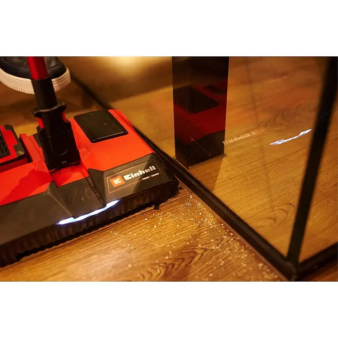 Einhell Seitenbürsten-Set - 4 Stk., Passend für: Einhell Akku-Besen TE-FS 18 Li - Ein roter Einhell Akku-Besen reinigt Krümel auf einem Holzboden neben einem schwarzen Tischbein unter einem Glassockel.