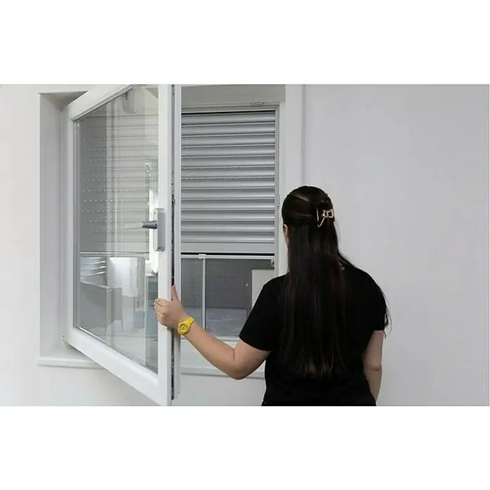 Schellenberg Mosquitera - 75 cm - 142 cm, Inserción, Color bastidor: Blanco - Una mujer está abriendo una ventana con una persiana enrollable, que está parcialmente bajada, en una habitación luminosa. La ventana es blanca y tiene un mango.