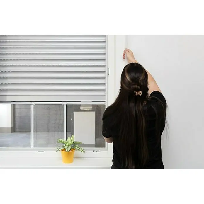 Schellenberg Mosquitera - 75 cm - 142 cm, Inserción, Color bastidor: Blanco - Mujer abriendo una ventana con persianas enrollables y mosquitera. Una planta en maceta se encuentra en el alféizar de la ventana.