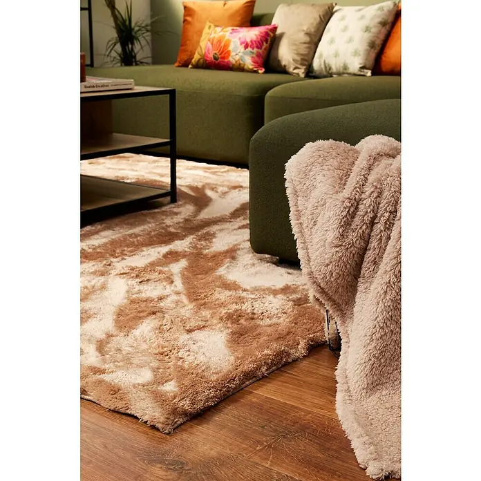 Alfombra de pelo largo Cuba - Beige, 170 x 120 cm, 100% Poliéster - Primer plano de una alfombra shaggy beige con un patrón irregular, colocada sobre un suelo de madera, junto a un sofá verde y un plaid beige.