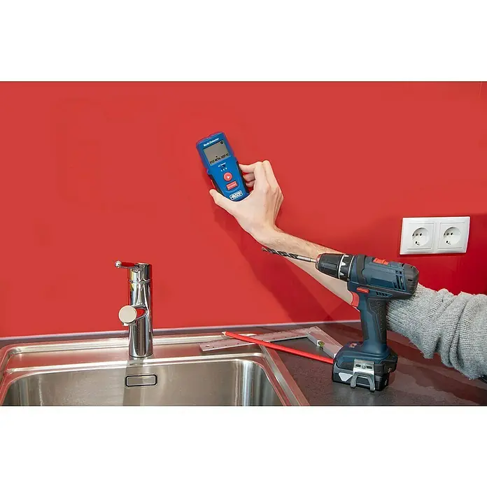 Alpha Tools Metall- & Holzsuchgerät Multi-Detektor - Betriebstemperatur: 0 °C - 40 °C - Nahaufnahme eines blauen Multi-Detektors, der an einer roten Wand gehalten wird, während eine Bohrmaschine und eine Steckdose in der Nähe zu sehen sind.