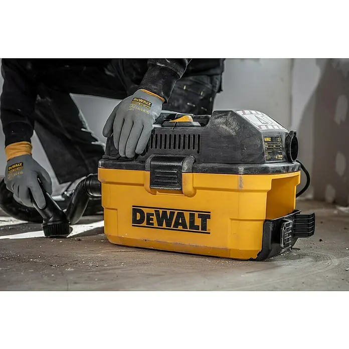 DeWalt Elektro-Nass- & Trockensauger DXV23G - 230 W, Behältervolumen: 23 l - Ein gelber und schwarzer DeWalt Staubsauger, der auf einem staubigen Boden steht. Der Arbeiter benutzt ihn, um den Boden zu reinigen.