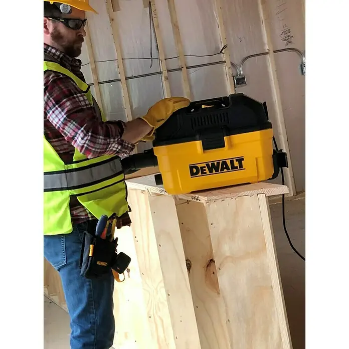 DeWalt Elektro-Nass- & Trockensauger DXV15T - 1.100 W, Behältervolumen: 15 l, Filterreinigung: Vollautomatisch - Nahaufnahme eines gelben DEWALT-Staubsaugers, der auf einem Holzbock steht, wobei ein Bauarbeiter im Hintergrund steht.