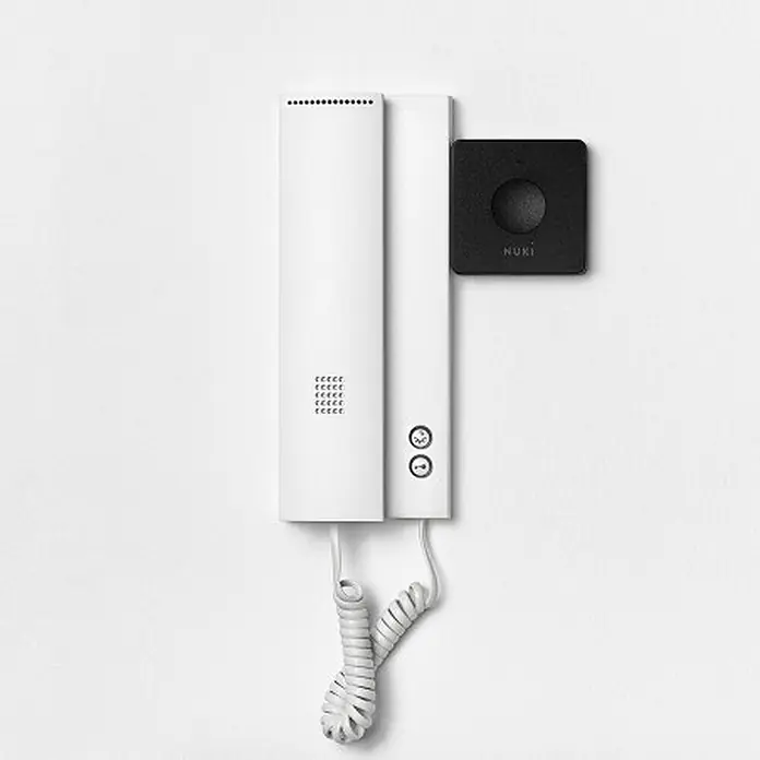 Nuki Control de apertura de puertas Opener - L x An x Al: 25 x 70 x 70 mm, WLAN - La imagen muestra un intercomunicador HUKI blanco con un cable en espiral montado en una pared blanca, acompañado de un accesorio negro cuadrado.