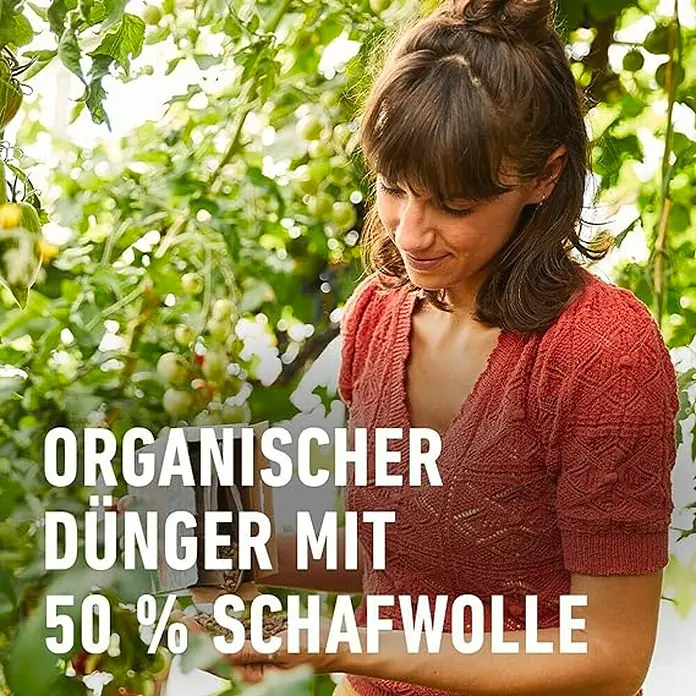 Compo Bio-Schafwolldünger Tomaten - 0,75 kg, Gemüse - Eine Frau in einem Gewächshaus, die biologischen Dünger mit 50% Schafwolle bewirbt. Der Fokus liegt auf dem Produkt inmitten der Pflanzen.