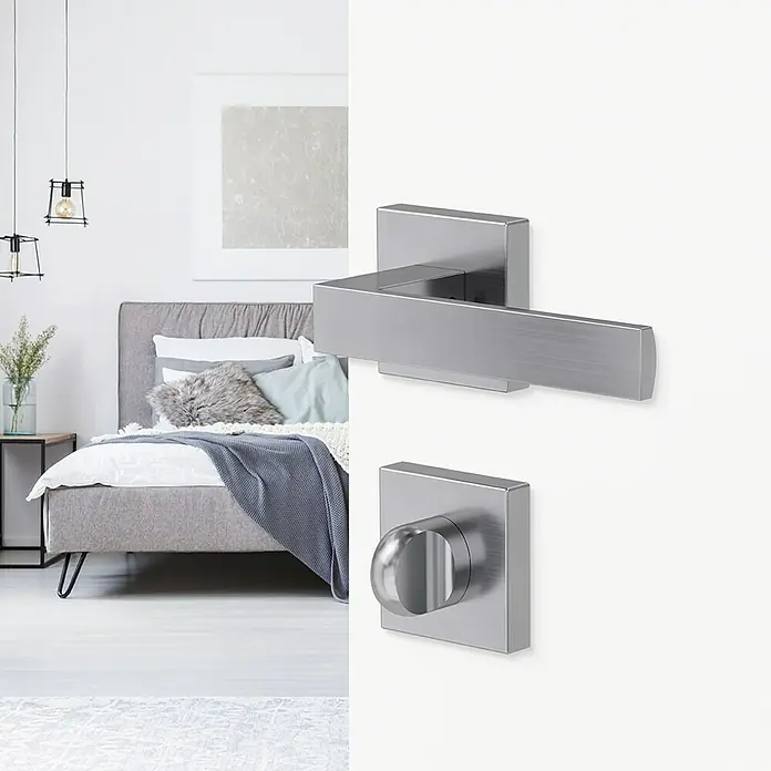 Diamond Doors Creative Zimmertürgarnitur WC Savannah - Stärke Tür: 40 mm - 45 mm, Edelstahl-Optik matt, Schlitzkopf/Olive - Nahaufnahme einer modernen Türschnalle aus gebürstetem Edelstahl mit einem quadratischen Design vor einem Schlafzimmerhintergrund.