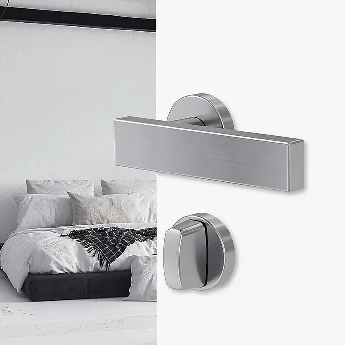 Diamond Doors Modern Zimmertürgarnitur WC Cube - Edelstahl, Schlitzkopf/Olive - Nahaufnahme eines modernen Türgriffs aus gebürstetem Stahl vor einem Schlafzimmerhintergrund.