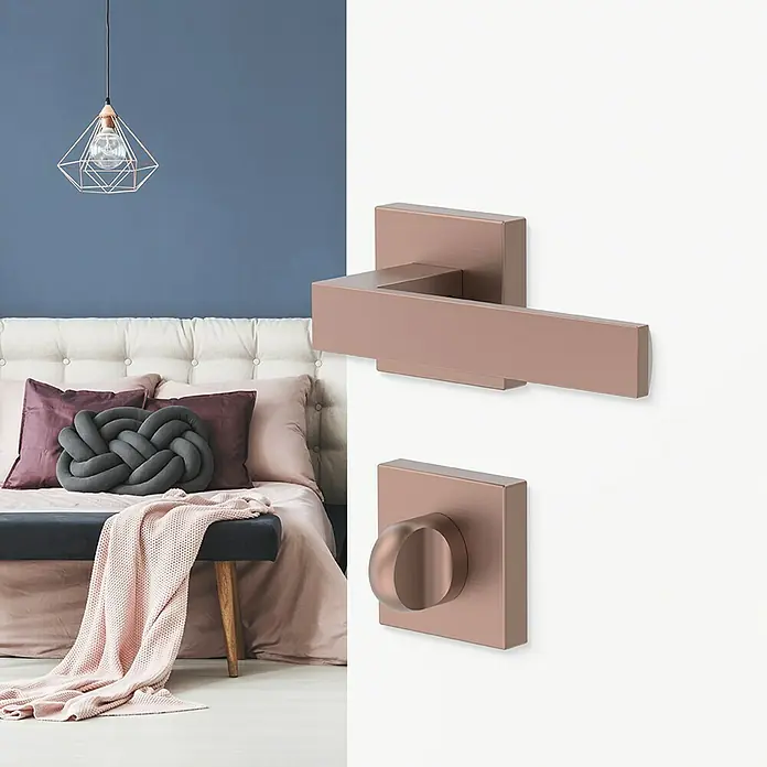 Diamond Doors Creative Zimmertürgarnitur WC Savannah - Stärke Tür: 40 mm - 45 mm, Kupfer, Schlitzkopf/Olive - Nahaufnahme eines modernen Türgriffs und eines Drehriegels in Kupferoptik vor einem Schlafzimmerhintergrund.
