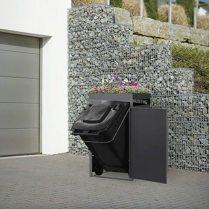 ML-Design Mülltonnenbox Mülltonnencontainer mit Pflanzdach für 1 Tonne 120 oder 240 Liter - Stahl, Eiche hell, Geeignet für Anzahl Mülltonnen: 1 Stk. - Eine graue Mülltonnenbox mit Bepflanzung, die eine schwarze Mülltonne versteckt, steht neben einer Garage. Ideal für die stilvolle Müllentsorgung.