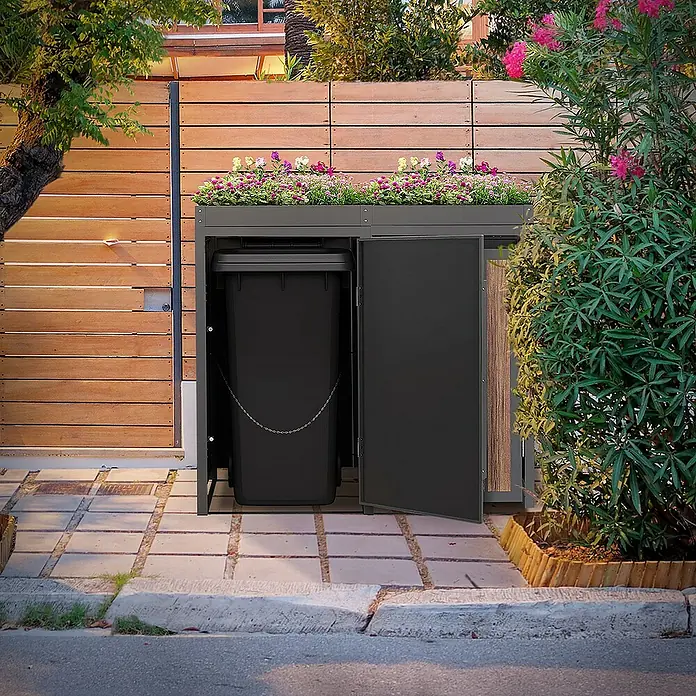 ML-Design Mülltonnenbox Mülltonnencontainer mit Pflanzdach für 2 Tonnen 120 oder 240 Liter - Stahl, Eiche dunkel, Geeignet für Anzahl Mülltonnen: 3 Stk. - Mülltonnenbox aus anthrazitfarbenem Metall mit Pflanzkasten auf Betonsteinen und einem Holzschuppen im Hintergrund.